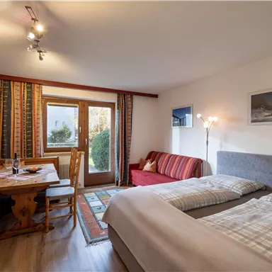 Ein gemütliches Zimmer mit zwei Betten, einem Essbereich und einem Sofabereich. Durch die Fenster fällt Licht und es gibt eine schöne Aussicht ins Freie.