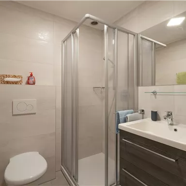 Ein modernes Badezimmer mit einer Dusche und einem Waschbecken. Die Wände sind hell gefliest, und es gibt Handtücher sowie einige Pflegeprodukte.