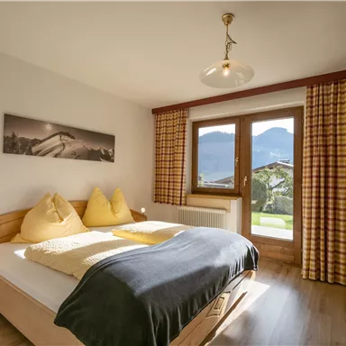 Ein gemütliches Schlafzimmer mit einem Doppelbett und zwei Dekokissen. Große Fenster bieten einen Blick auf die Berge und viel natürliches Licht.