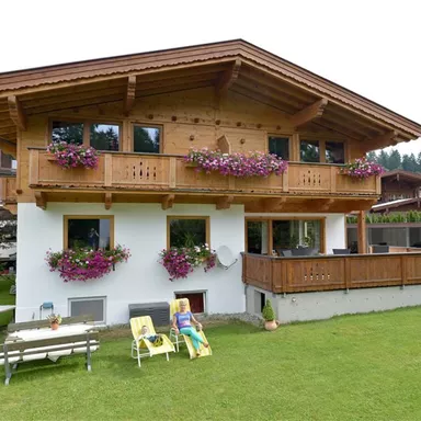 A cozy, two-story wooden house with blooming flower boxes. The garden is green and offers loungers for relaxation.