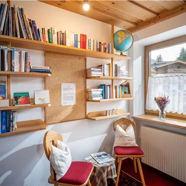 A cozy reading nook with a bookshelf on the wall. Two chairs and a small table are positioned in front of a window with a view outside.