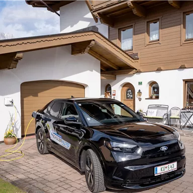 A black car is parked in front of a traditional, wooden-clad house. In the background, some garden furniture and a clear sky are visible.