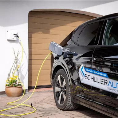A black electric car is being charged at a charging station. In the background, a garage can be seen.