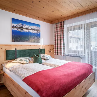 A cozy bedroom with a large bed and wooden decor. The room has a balcony and a beautiful picture of mountains on the wall.