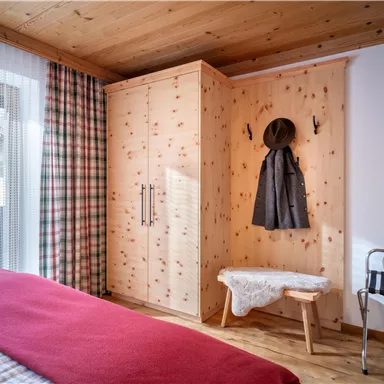 A cozy bedroom with wooden walls and a large wardrobe. On a bed lies a red blanket and beside it stands a bench with a hat.