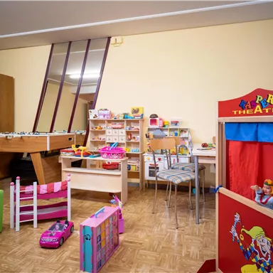 A colorful play area with various toys, including a puppet theater and a pool table. The walls are bright, and the room feels welcoming for children.