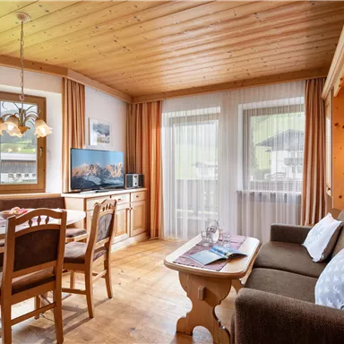 A cozy living space with wooden ceilings and large windows. There is a dining table, a television, and a seating area with a sofa.