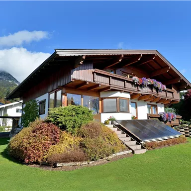 Ein charmantes Holzhaus mit Balkon und bunten Blumen. Es liegt in einer grünen Umgebung mit niedrigen Bergen im Hintergrund.