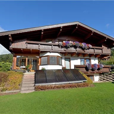Ein schönes, traditionelles Holzhaus mit einem großen Garten. Auf dem Balkon hängen bunte Blumenkästen und die Sonne scheint.