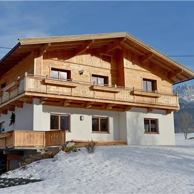A cozy wooden house with a balcony, surrounded by snow-covered ground. Gentle mountains are visible in the background.