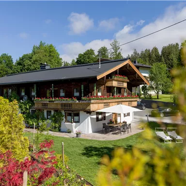 A beautiful house in the greenery with a large garden. On the terrace, there are furniture and flowers are blooming in the surrounding area.