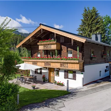 A charming wooden house with balconies and blooming floral decorations. In the foreground, there is a terrace and in the background, trees and a blue sky can be seen.