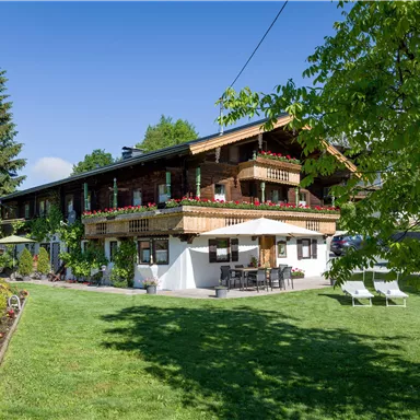 A charming wooden house with green outdoor areas and a beautiful garden. The terrace is equipped with seating, and there are many plants in the foreground.