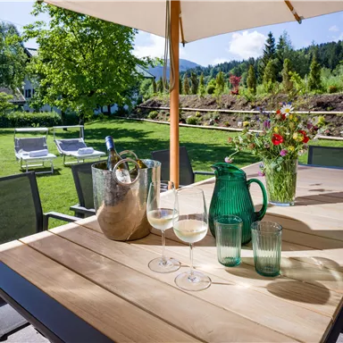 A cozy table in the garden, decorated with glasses, a carafe, and a vase of flowers. In the background, green trees and a well-kept garden can be seen.