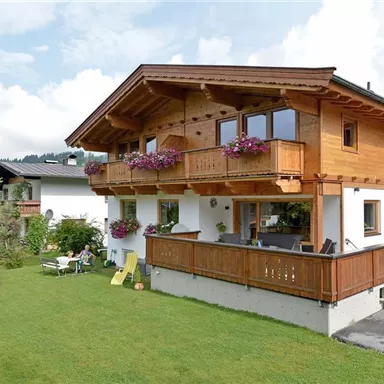 A charming wooden house with a balcony and colorful flowers. The garden is green and inviting, perfect for relaxing.