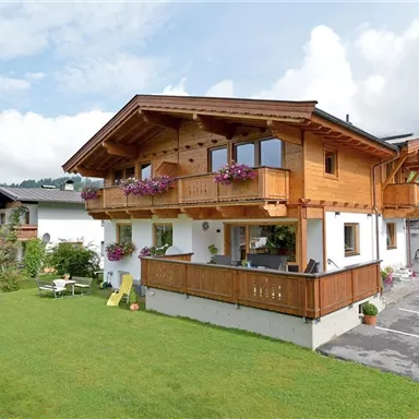 A charming holiday home in alpine style with balconies and colorful flowers. Surrounded by a green meadow and mountains in the background.