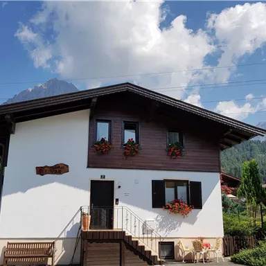 A charming house with a white part and a wooden-paneled part. Surrounded by mountains and a green garden.
