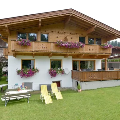 A cozy wooden house with a balcony, decorated with colorful flowers. In the foreground, there are deck chairs and a table on a green meadow.