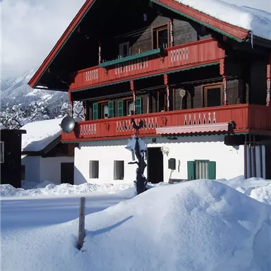 A traditional wooden house with red balcony decorations, surrounded by snow. In the background, mountains and a clear sky can be seen.