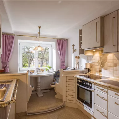 Eine moderne Küche mit hellem Holzdesign und weißen Fliesen. Ein Tisch mit Stühlen steht vor einem Fenster, das viel Tageslicht hereinscheinen lässt.