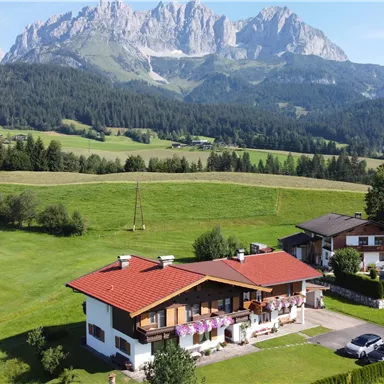 Eine malerische Landschaft mit grünen Wiesen und hohen Bergen im Hintergrund. Im Vordergrund befinden sich zwei gemütliche Häuser mit roten Dächern.