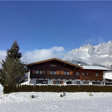Ein hübsches Holzhaus im Winter mit schneebedecktem Boden. Im Hintergrund sind verschneite Berge und ein blauer Himmel zu sehen.