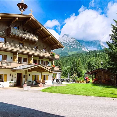 Ein traditionelles Alpenhaus mit mehreren Balkonen und schönen Blumen. Im Hintergrund sind grüne Wiesen und Berge zu sehen.