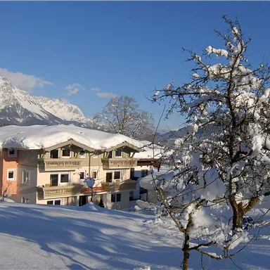 A cozy house in the snow with a bright blue sky. The surroundings are characterized by snow-covered mountains and trees.