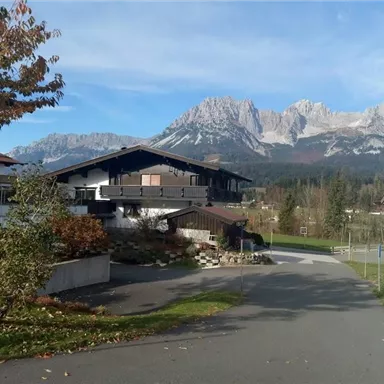 A picturesque residential area with modern houses and a view of impressive mountains in the background. The sky is clear, and the surroundings are green and inviting.
