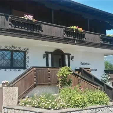 A cozy chalet with a wooden balcony and blooming plants. In the background, green meadows and high mountains can be seen.