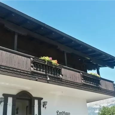 A traditional chalet with a wooden balcony and colorful flower boxes. In the background, majestic mountains and a clear blue sky can be seen.