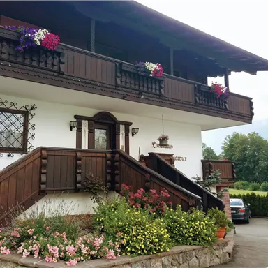 A traditional alpine house with a wooden balcony decorated with flowers. In front of the house, colorful flowers and parked cars can be seen.