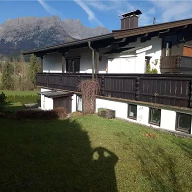 A modern house with a large garden and a view of the mountains. The surroundings are green and surrounded by nature.