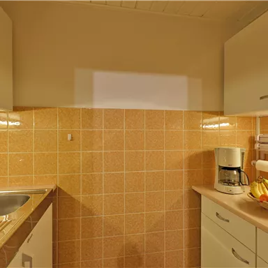 A small kitchen with bright tiles and modern furniture. On the countertop, there is a coffee machine and a bowl of fresh fruit.