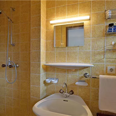 A small bathroom with a shower cabin and a sink. The walls are tiled with yellow tiles and there is a mirror above the sink.