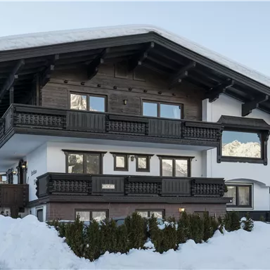 Ein imposantes Chalet in den Alpen, umgeben von Schnee. Das Gebäude hat einen traditionellen Holzstil mit Balkonen und großen Fenstern.