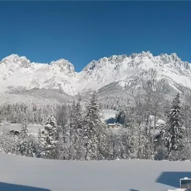 A winter landscape with snow-covered mountains and clear blue sky. In the foreground, there are snow-covered trees and a calm atmosphere.