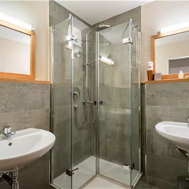 Ein modernes Badezimmer mit Glasdusche und zwei Waschbecken. Die Wände sind mit grauen Fliesen verkleidet und es gibt Holzdetails.