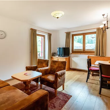 Eine gemütliche Wohnstube mit braunen Sesseln und einem Tisch. Große Fenster bringen viel Licht in den Raum.