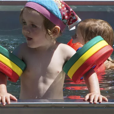 Ein kleines Kind mit bunten Schwimmflügeln steht am Rand eines Schwimmbeckens. Im Hintergrund sind weitere Kinder im Wasser zu sehen.