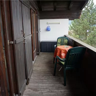 A balcony with two chairs and a table covered with an orange tablecloth. In the background, trees and a mountain landscape are visible.