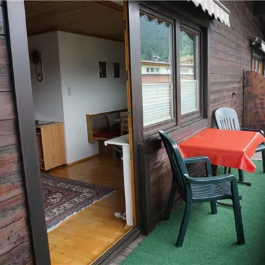 A cozy balcony with a red table and chairs. In the background, there is an open door to a wood interior.