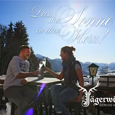 Ein Paar sitzt an einem Tisch in den Bergen und genießt ein Getränk in der Sonne. Im Hintergrund sind verschneite Berge und Tannenbäume zu sehen.