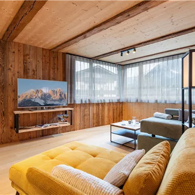 A modern living room with wood paneling and large windows. On the table is a cozy yellow couch and a TV with a mountain view.