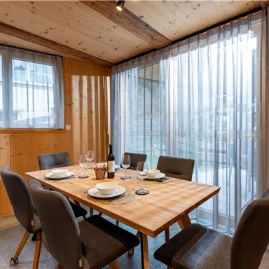 A cozy dining area with a wooden tray and elegant chairs. Large windows with curtains let in plenty of light.