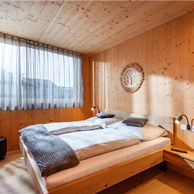 A modern bedroom with wooden walls and a large window. The room is bright and cozy with a double bed and a TV corner.