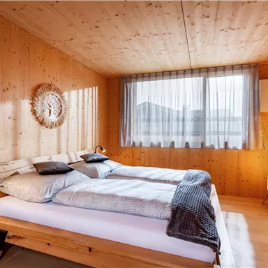 A modernly furnished bedroom with wood paneling and large windows. It features two single beds, cozy lighting, and a comfortable style.