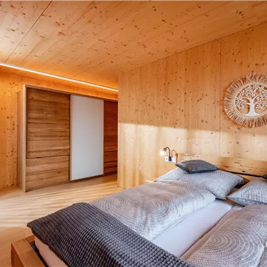 A modern bedroom with wood walls and a large window. The bed is cozy, decorated with gray pillows and blankets.