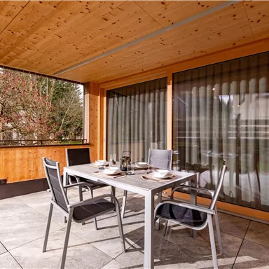 A cozy dining area on a terrace with a table and chairs. The windows are fitted with curtains, and the room feels inviting.