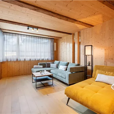 A bright living room with wooden walls and large windows. There are comfortable sofas in soft colors and a modern television.